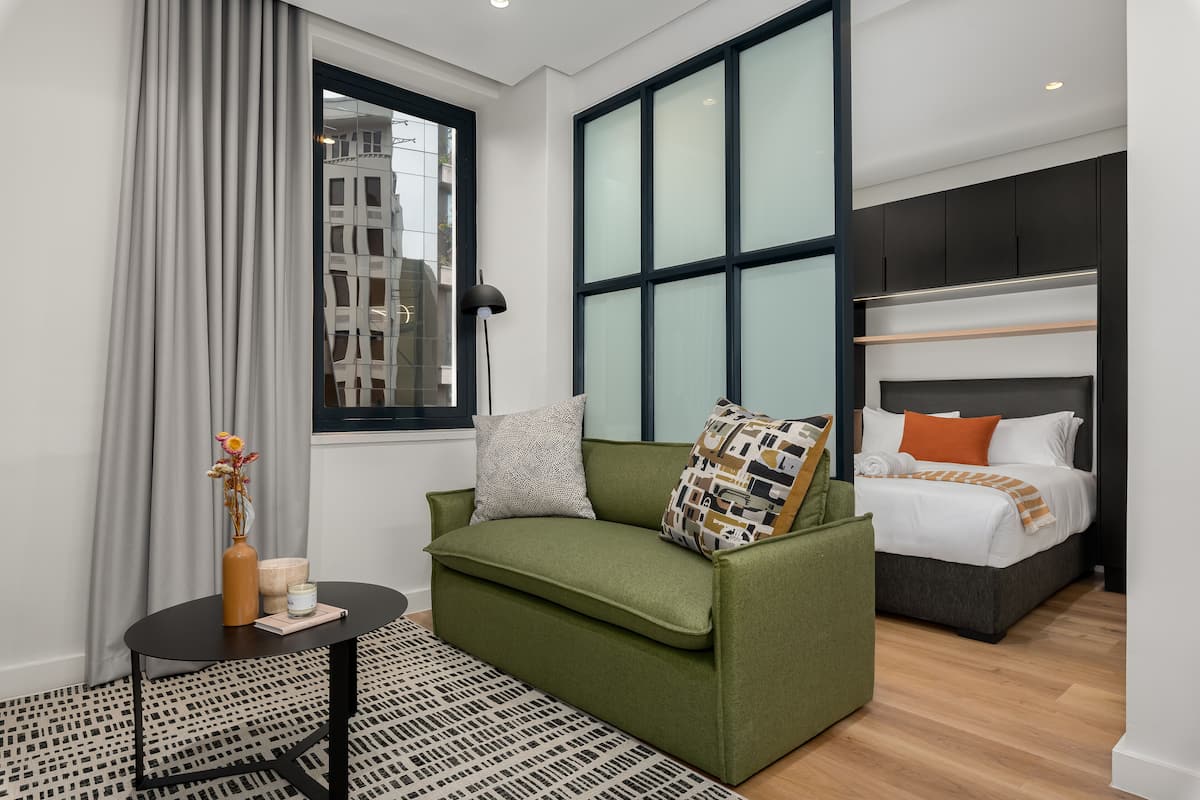 Una habitación de hotel moderna con un sofá verde, una pequeña mesa redonda, una cama con cabecera de madera y ventanas amplias con vista a edificios.