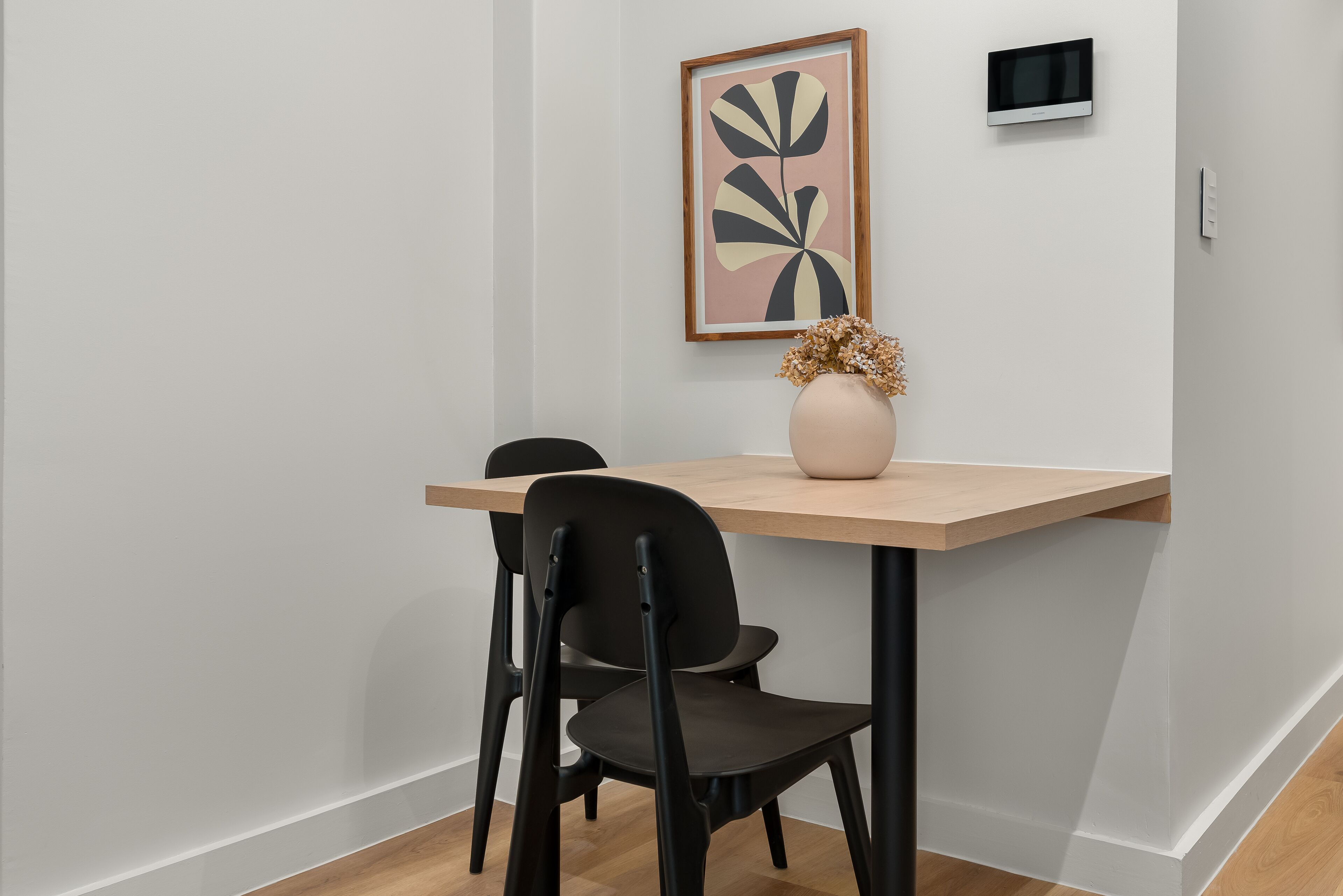 Ein Essbereich mit Holztisch, schwarzen Stühlen, einer Vase mit Trockenblumen und einem gerahmten Kunstwerk an der Wand.