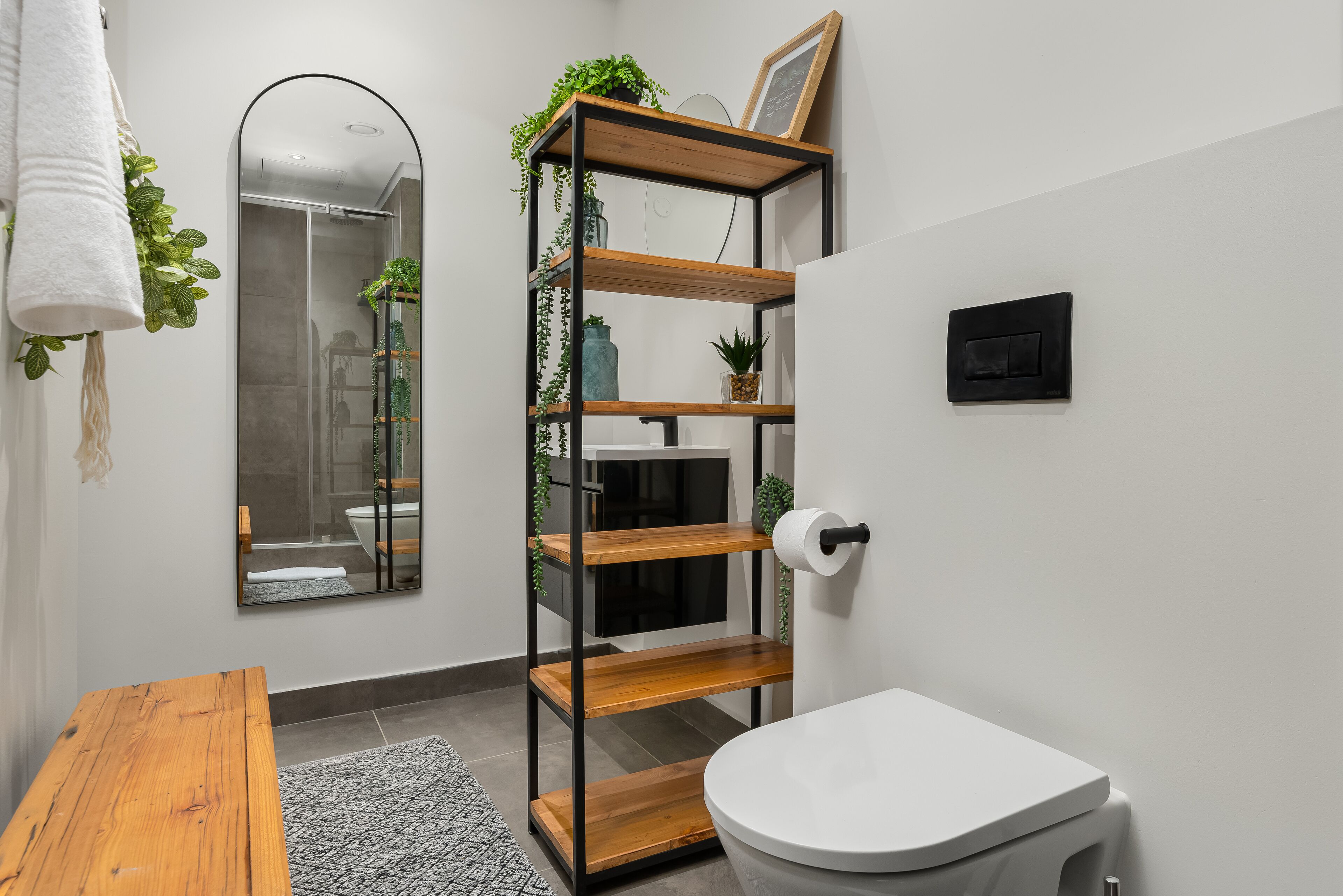 A bathroom with a white toilet, a wooden shelf with plants, a mirror, and a towel hanging on the wall.