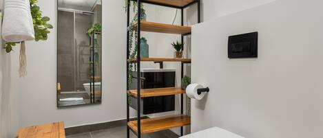 A bathroom with a white toilet, a wooden shelf with plants, a mirror, and a towel hanging on the wall.
