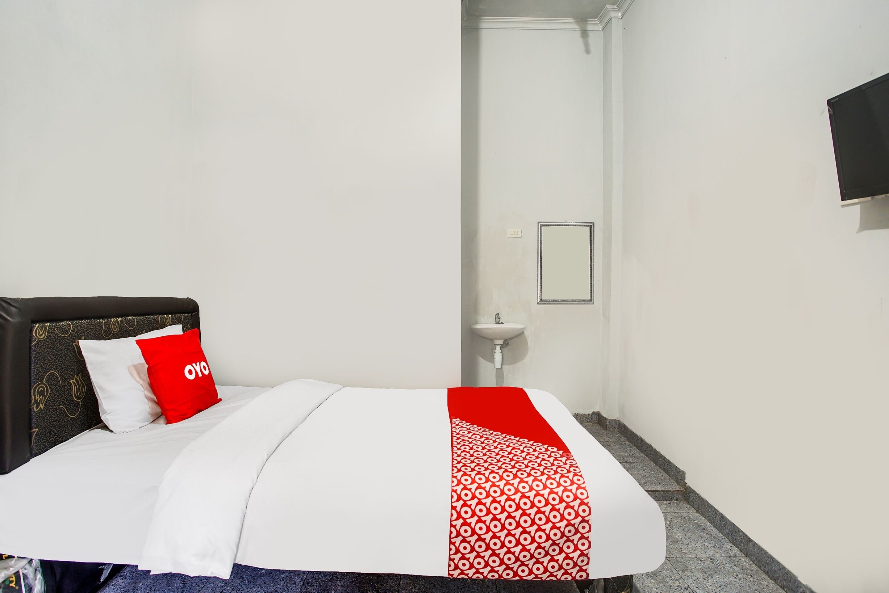 Habitación de hotel con cama, cubrecama rojo y blanco, almohada blanca, almohada roja, cabecero negro, pared blanca, lavamanos blanco y espejo.