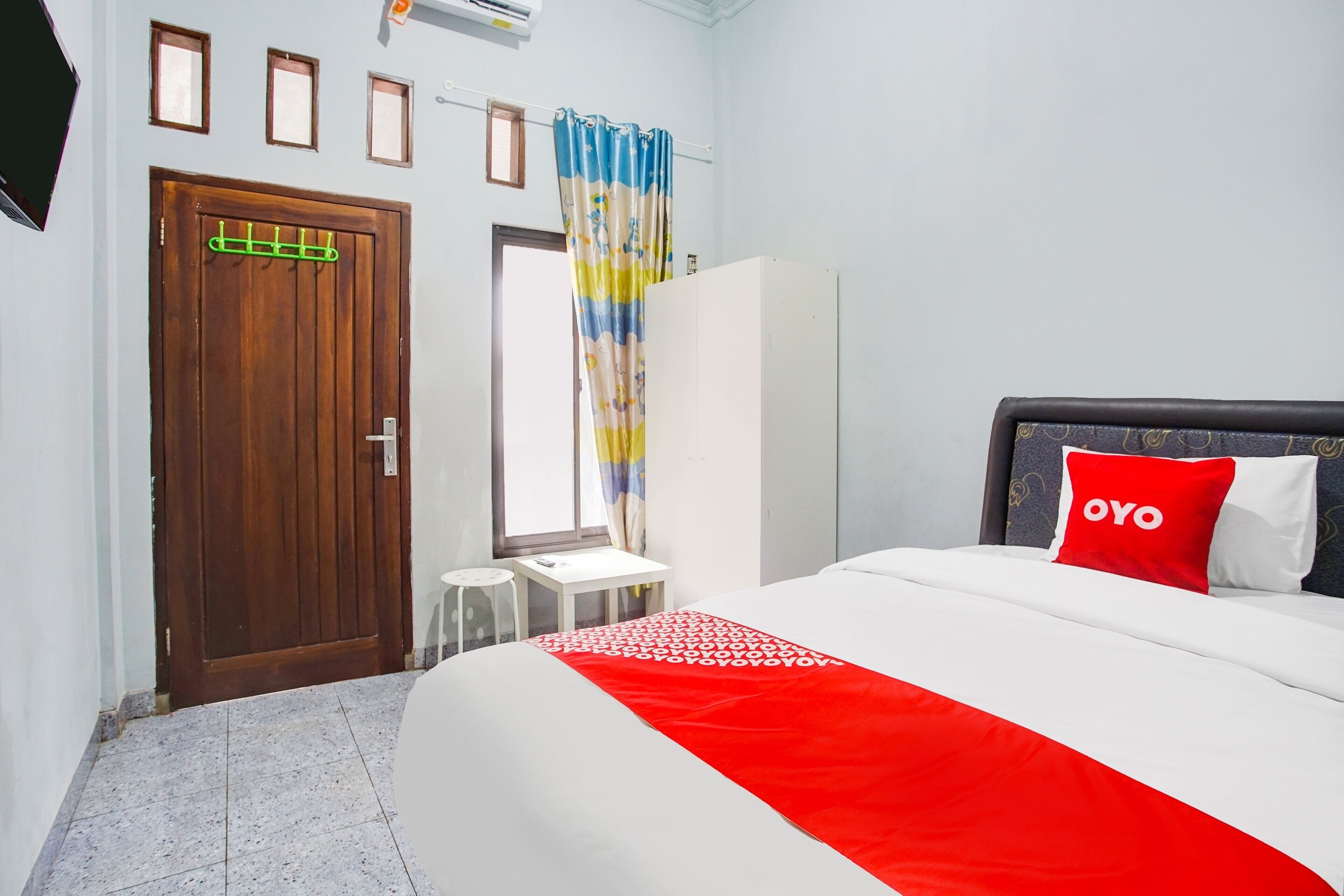 A hotel room with a wooden door, a small table, a bed with a red and white bedspread, and a window with curtains.