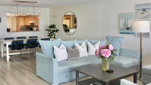 A modern living room with a light blue sofa, a coffee table, and a dining area with a white table and chairs. - Bayway Shores 303 - Waterfront Condo (CLEARWATER BEACH)