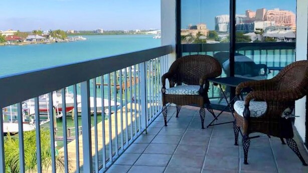 A balcony with two wicker chairs, a glass table, and a view of the sea and cityscape. - Bayway Shores 303 - Waterfront Condo (CLEARWATER BEACH)
