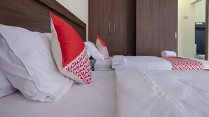 Quarto de hotel com duas camas, cada uma com roupa de cama branca e almofadas vermelhas e brancas. Possui uma cabeceira de madeira, um guarda-roupa com duas portas e uma toalha no chão.