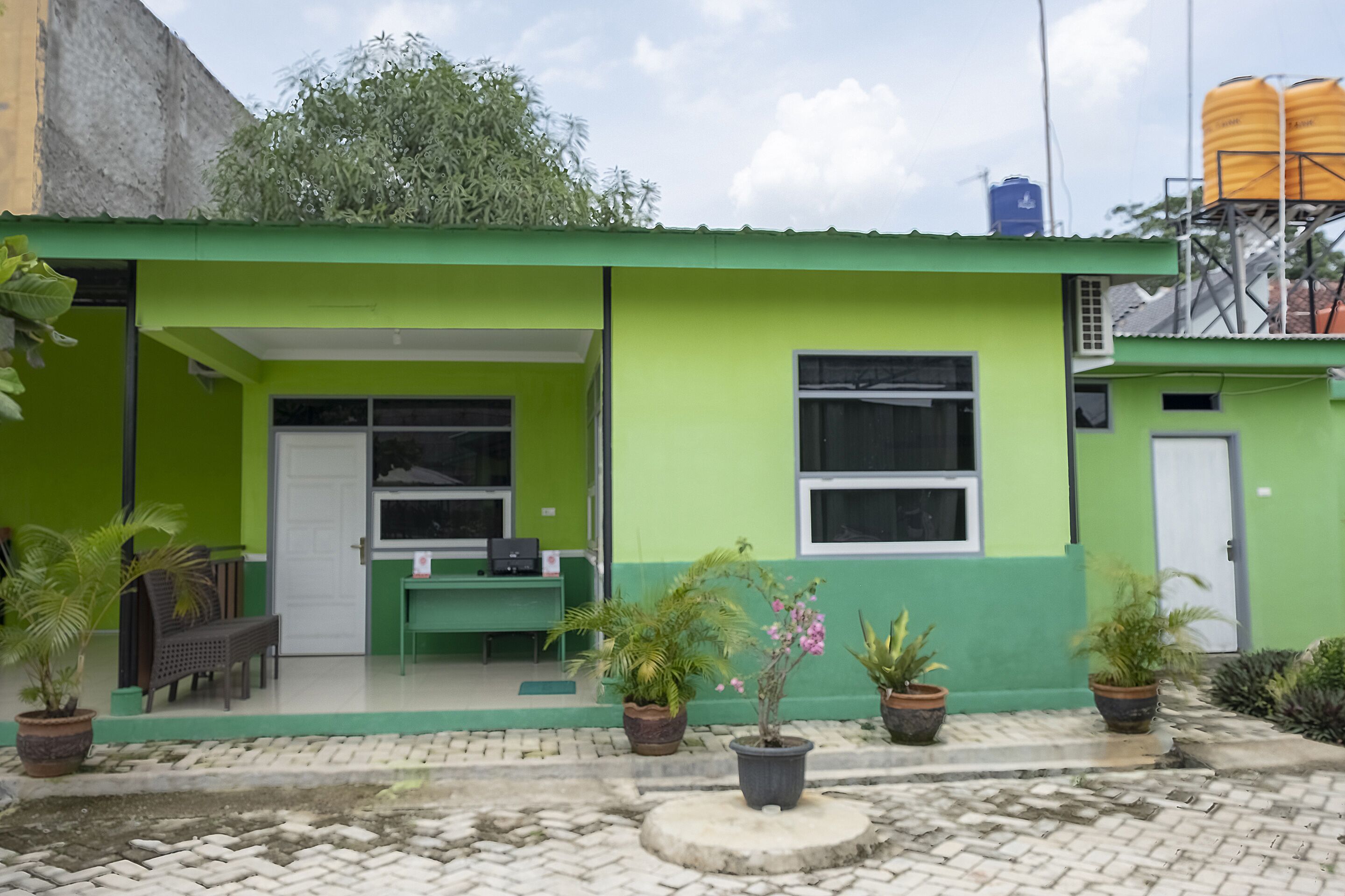 A green building with a covered patio area, potted plants, and a water tank on the roof.
