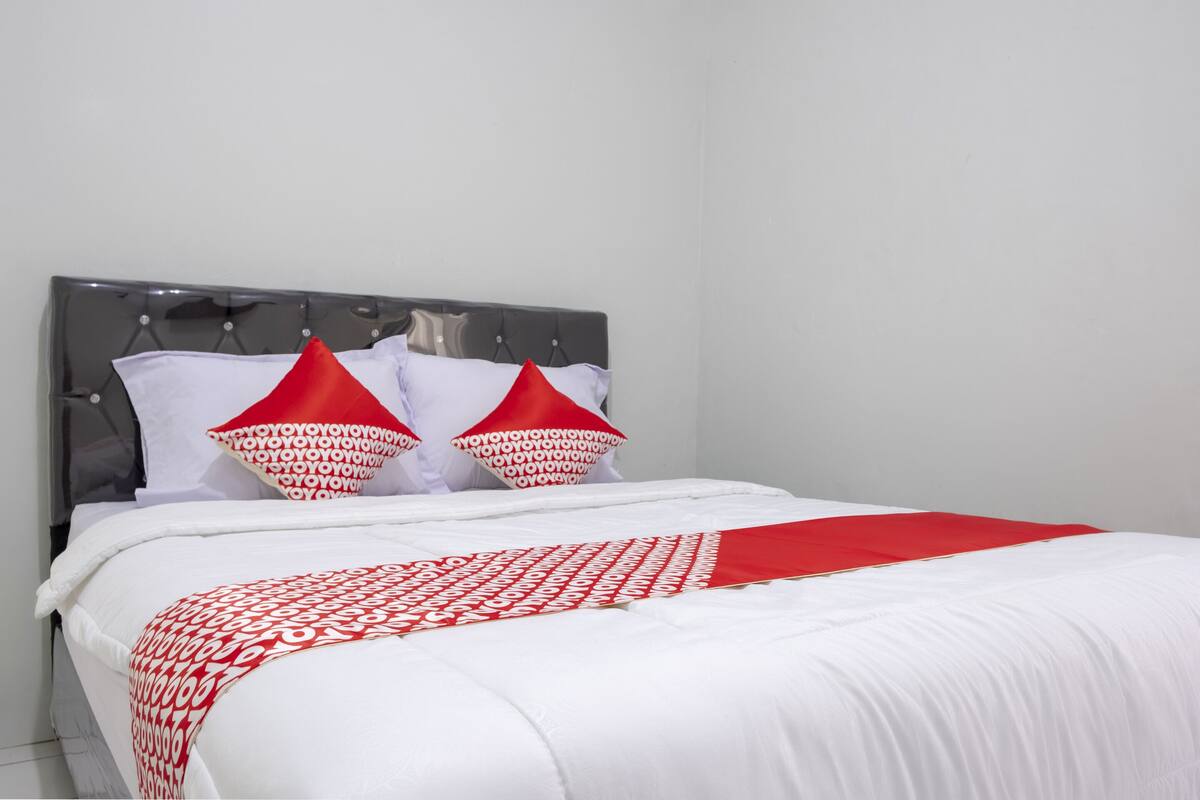 A neatly made bed with white bedding and red accents, a grey upholstered headboard, and two red pillows with white patterns.