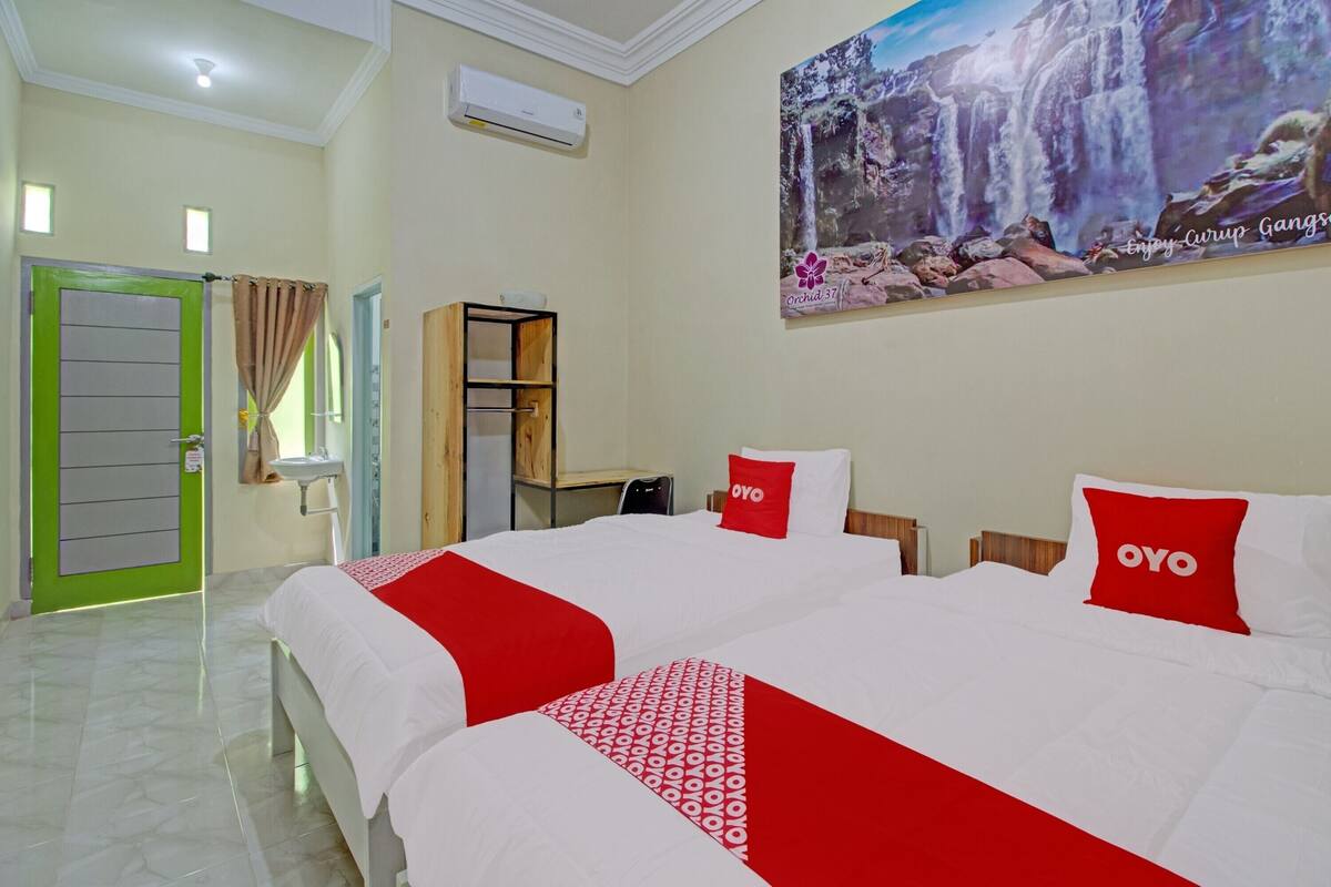 A hotel room with two beds, a green door, a white air conditioner, a wooden cabinet, and a large waterfall painting on the wall.