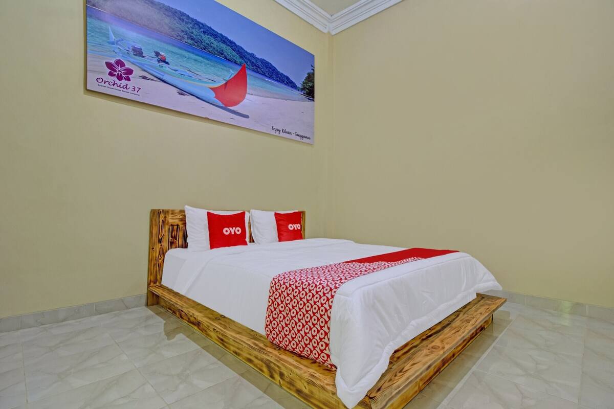A hotel room with a wooden bed frame, white bedding, and a red and white patterned blanket.