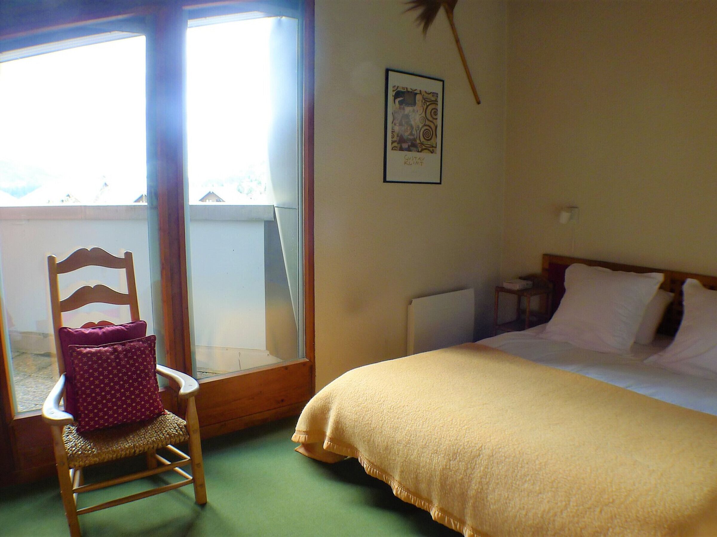 A bedroom with a bed, a chair with a cushion, a window with a view, and a framed picture on the wall.