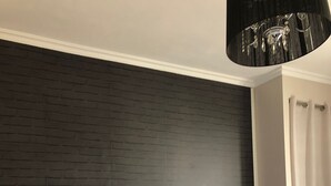 A bedroom with a bed, two bedside tables, a lamp on each table, a dark brick wall, and a black chandelier.