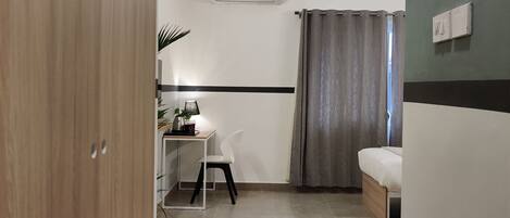 A compact room with a bed, a desk, a chair, a ceiling fan, and a window with curtains.