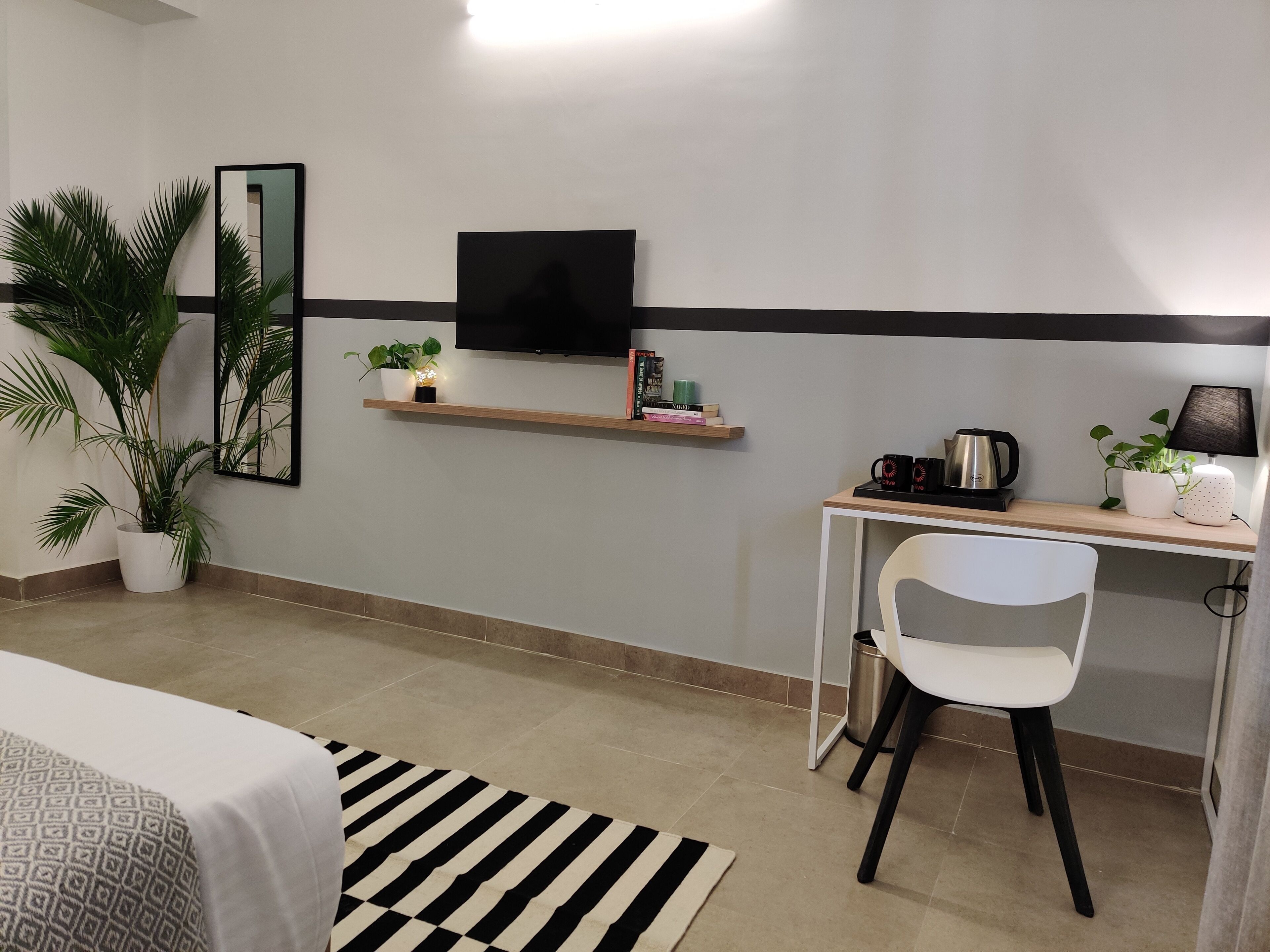 A hotel room with a bed, a desk with a chair, a TV, a shelf with books and a plant, and a potted plant beside the bed.