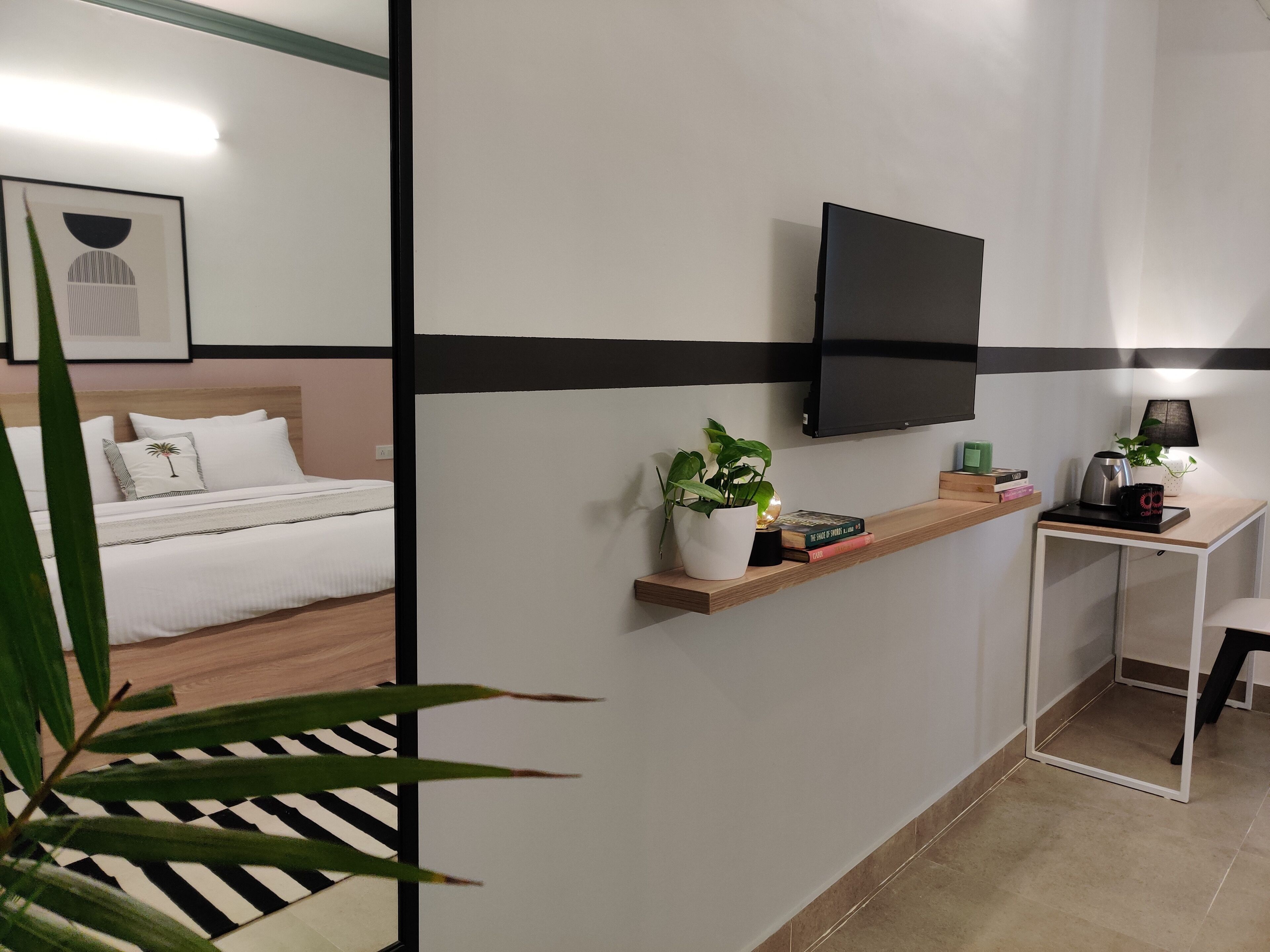 Una habitación de hotel moderna con una cama, un televisor montado en la pared, un escritorio pequeño con una lámpara y una estantería con libros y una planta.