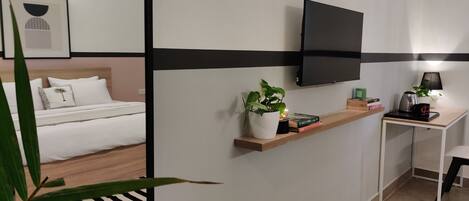 Una habitación de hotel moderna con una cama, un televisor montado en la pared, un escritorio pequeño con una lámpara y una estantería con libros y una planta.