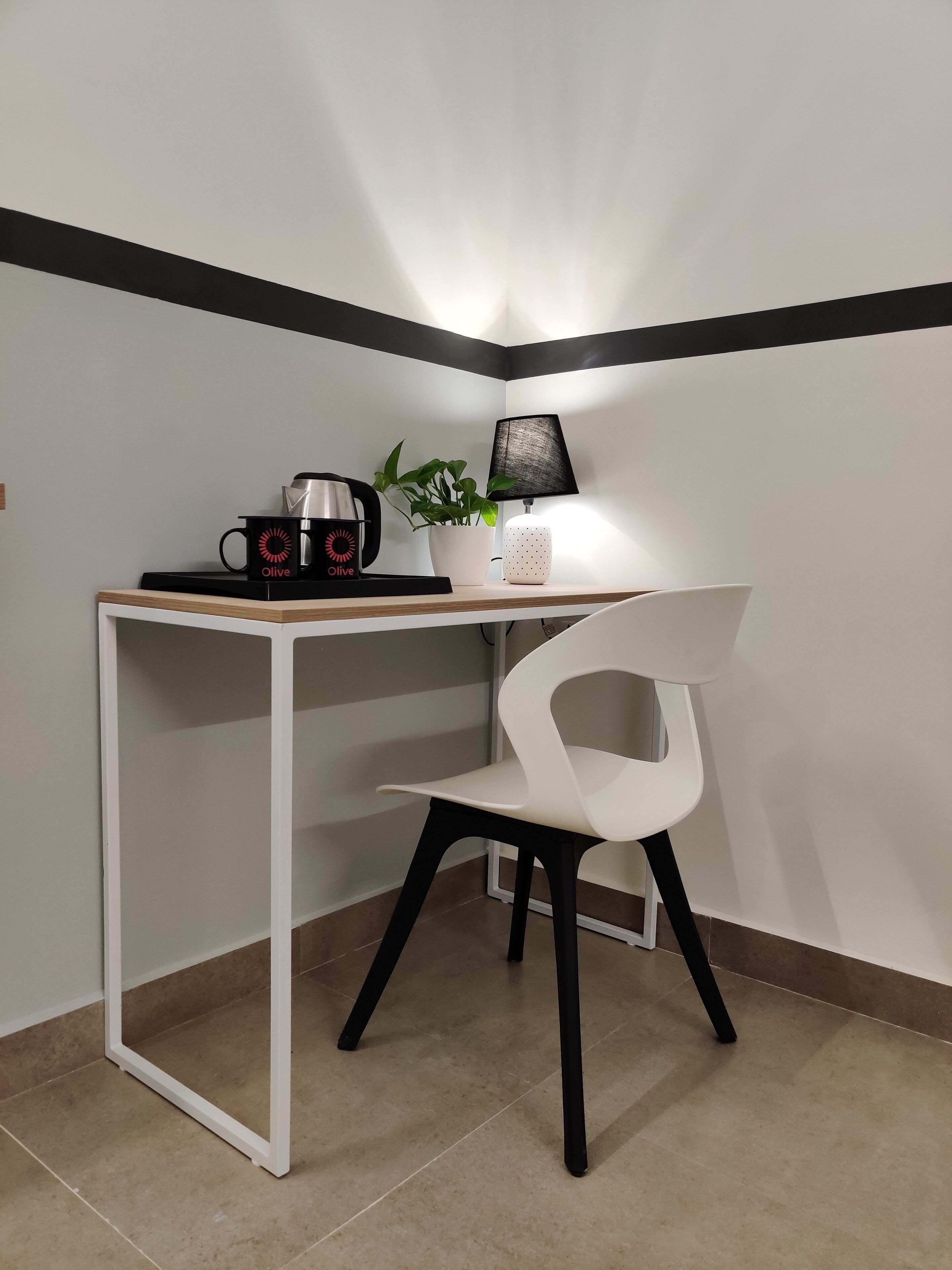 A small, white desk with a black and white striped wall, a white chair, a coffee maker, a potted plant, and a lamp.