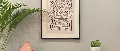 A framed abstract artwork on a wall, a potted plant, a pink vase, and a bookshelf with books and decorative items.