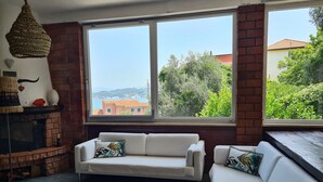 Un salon chaleureux avec un canapé blanc, un luminaire suspendu et une vue sur la verdure et les bâtiments, visible à travers de grandes fenêtres.