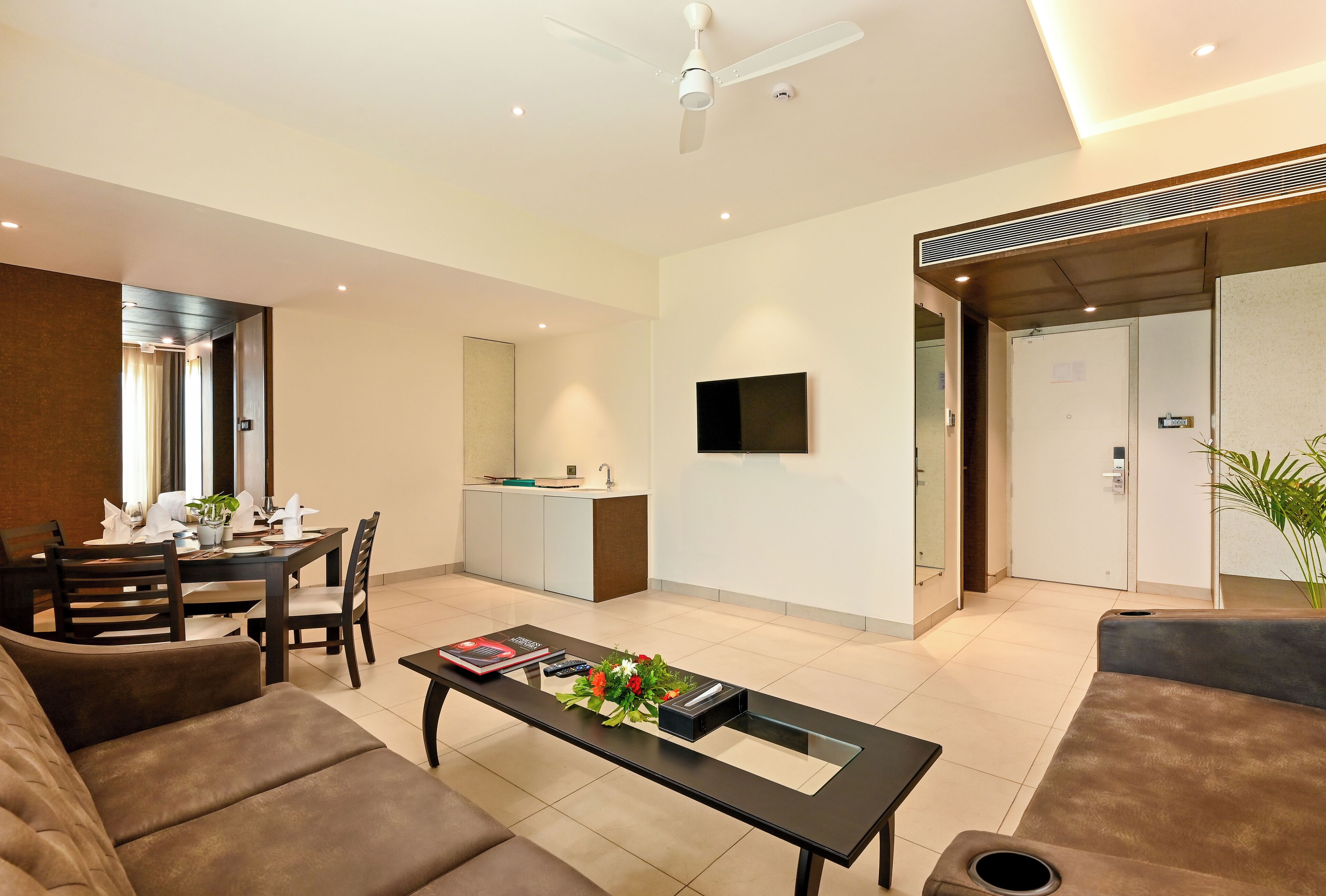Un salon moderne comprenant un canapé, une table basse avec des télécommandes et un arrangement floral, un coin repas avec des chaises et une table, un coin cuisine avec un évier et des placards, et un téléviseur fixé au mur.
