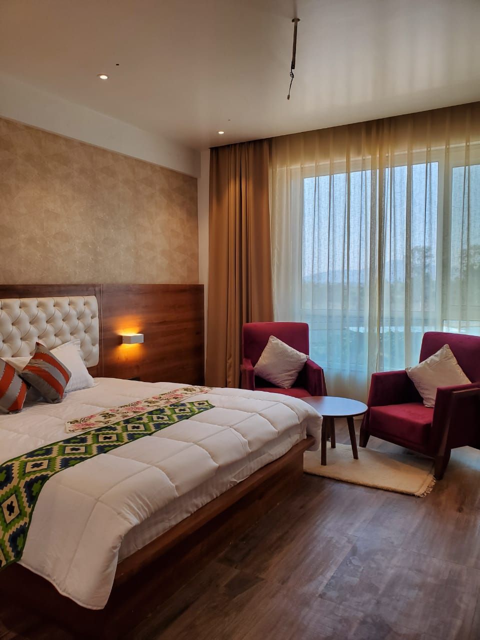 Habitación de hotel con una cama grande, dos sillones rojos, una mesita y vistas al exterior a través de cortinas traslúcidas.