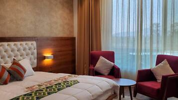 Habitación de hotel con una cama grande, dos sillones rojos, una mesita y vistas al exterior a través de cortinas traslúcidas.
