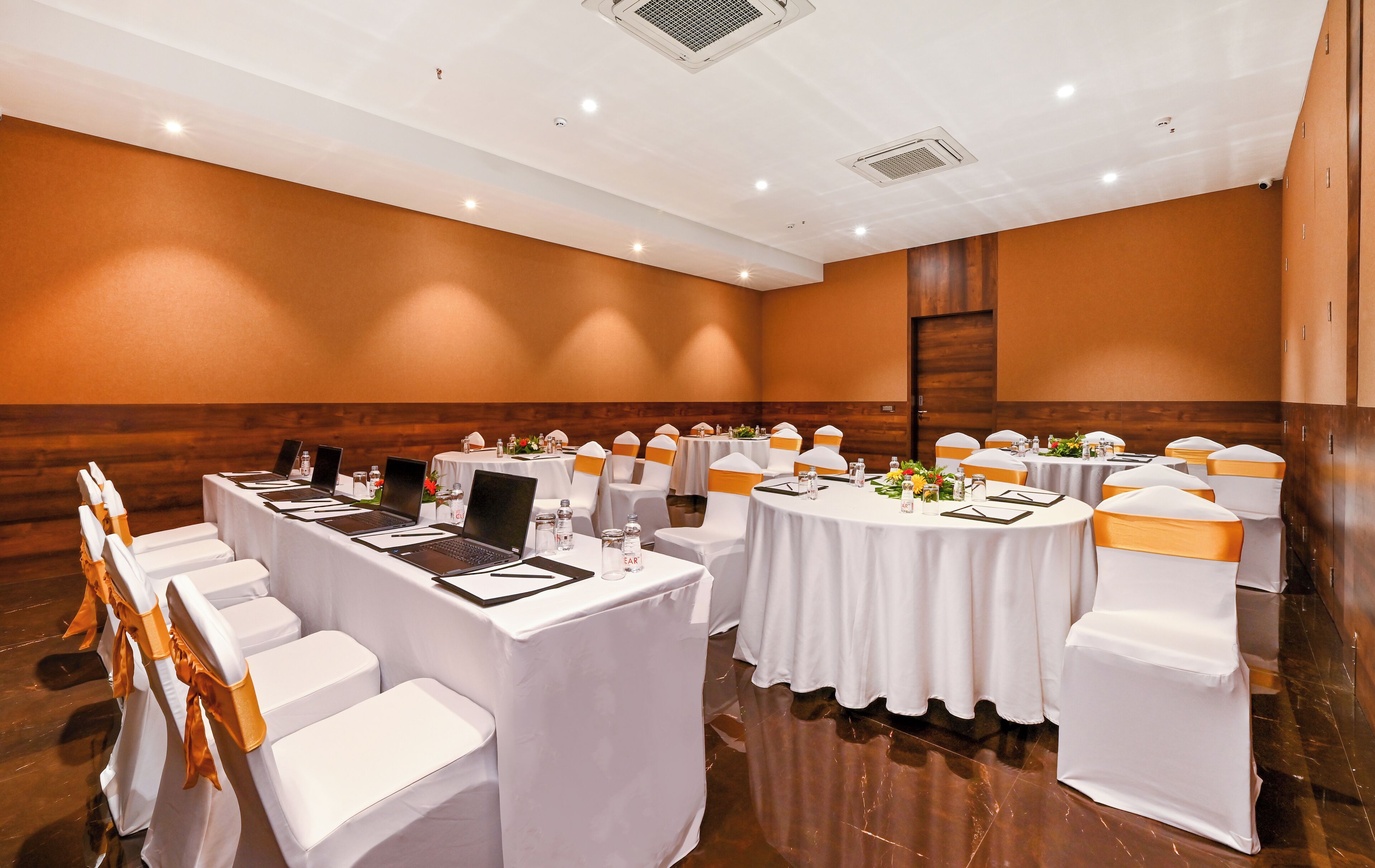 A conference room with round tables, white chairs with orange sashes, laptops, and water bottles.