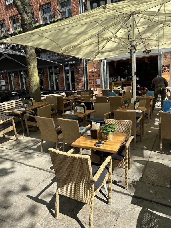 An outdoor cafe with wicker furniture, a large umbrella, and a brick building in the background.