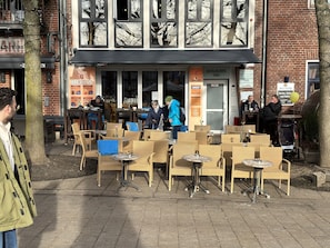 Un café en plein air avec des chaises et des tables en osier, un bâtiment en briques doté de grandes fenêtres et une enseigne indiquant « SCHLACHTE 30A ».
