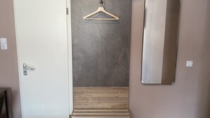 Een kamer met een witte deur, een houten bankje, een ophanghaak van hout en een grote spiegel aan de muur.