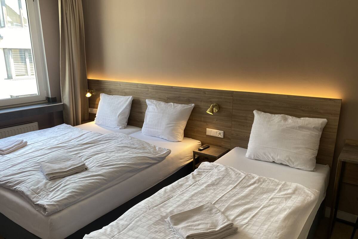 Hotelkamer met twee bedden, elk voorzien van witte lakens en kussens, een nachtkastje met een lamp, en een raam met gordijnen.