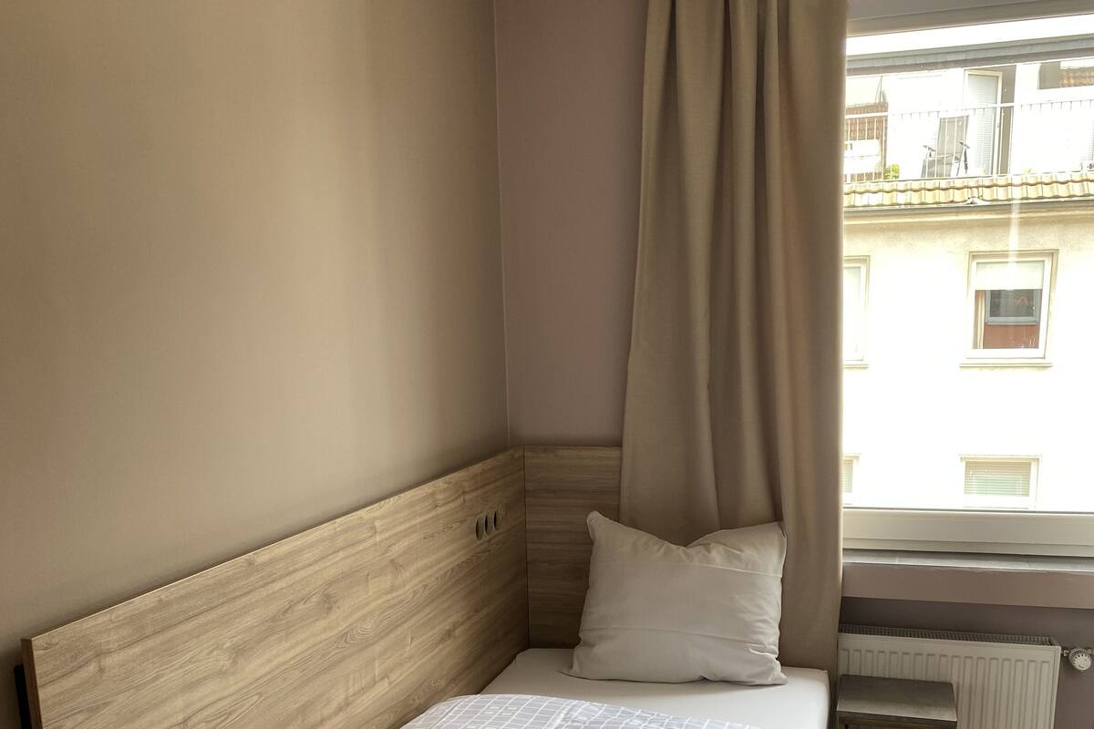 Een netjes opgemaakt bed met een houten hoofdbord, een klein nachtkastje en een raam met uitzicht op gebouwen.