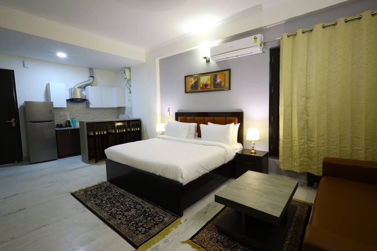 A hotel room with a bed, bedside table, and a small desk. There is a kitchenette with a refrigerator and a stove.