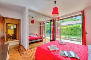 A hotel room with a large bed, a desk, a chair, and a view of greenery through the glass door.