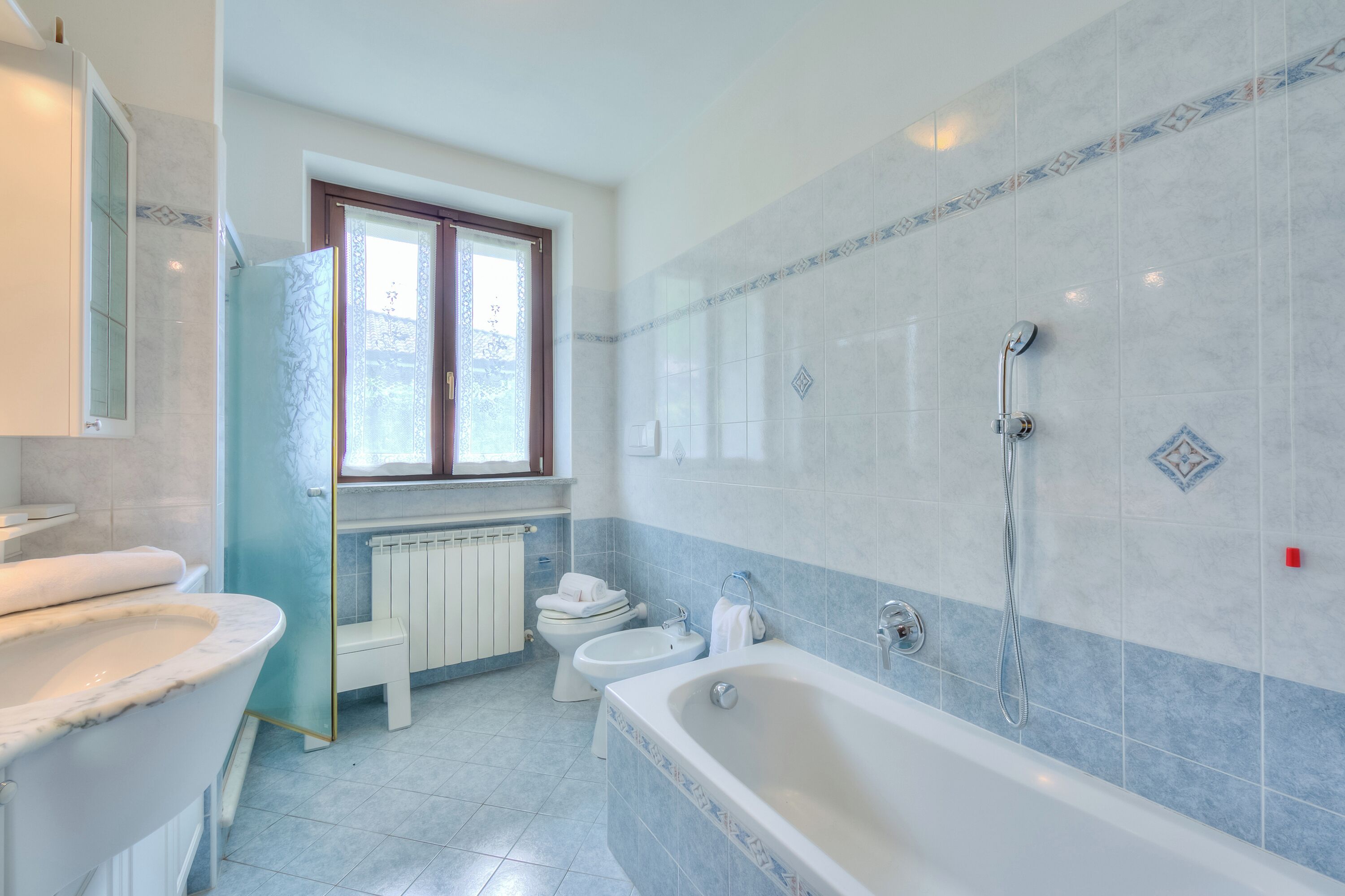 A bathroom with a bathtub, two toilets, a bidet, a glass shower enclosure, a window, and a radiator.