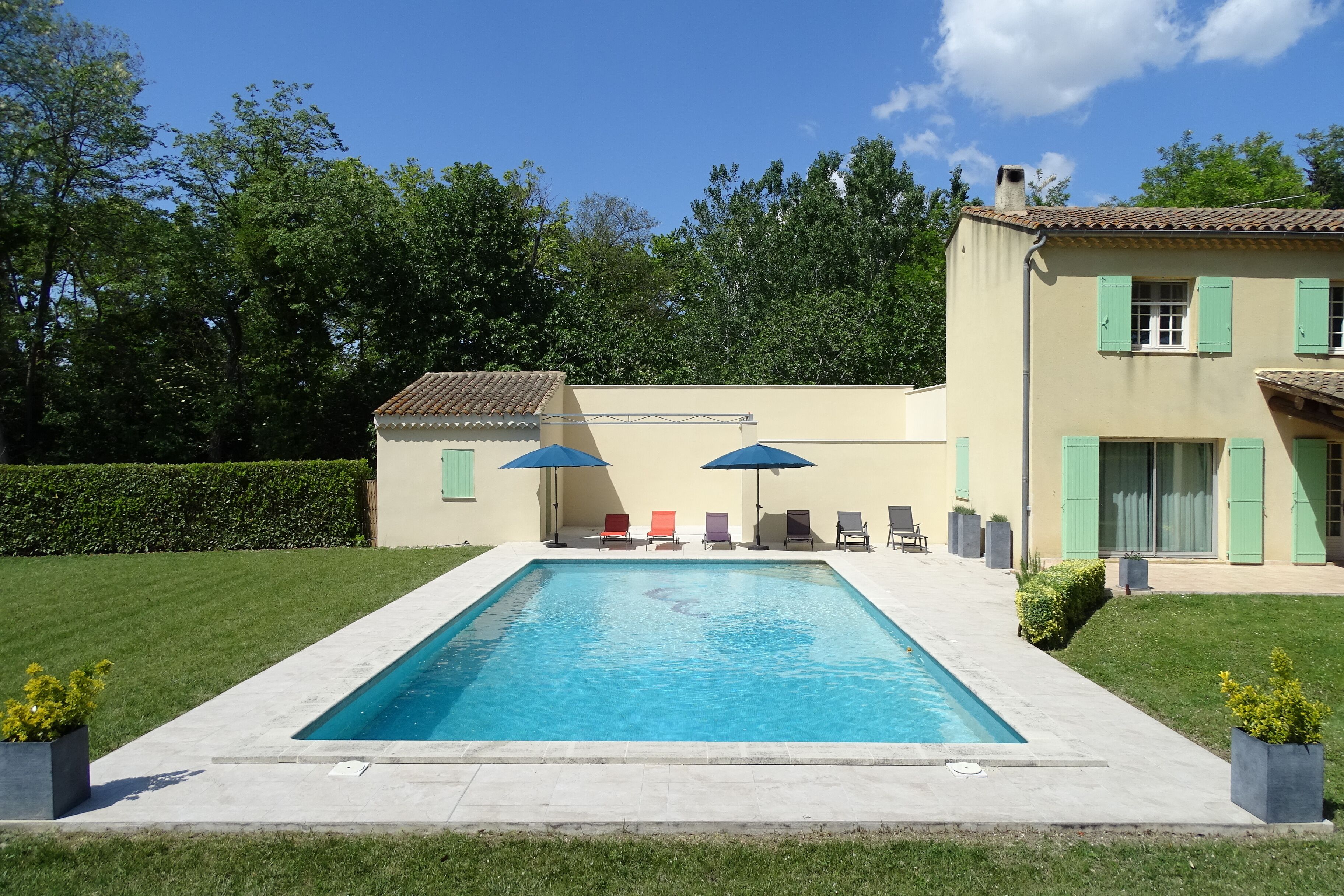 Una piscina rettangolare con bordo piastrellato, circondata da un prato curato e un edificio con persiane verdi.