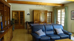 A living room with a blue leather sofa, wooden cabinets, a dining table with chairs, and a window with green curtains. - Villa "La Verdoulento" au Calme et à L'abri des Regards Avec Piscine 6mx12m (AVIGNON)