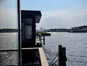 Blick auf das Wasser mit einer anlegenden Mole, einem Gebäude mit schrägem Dach und Industriegebäuden im Hintergrund.