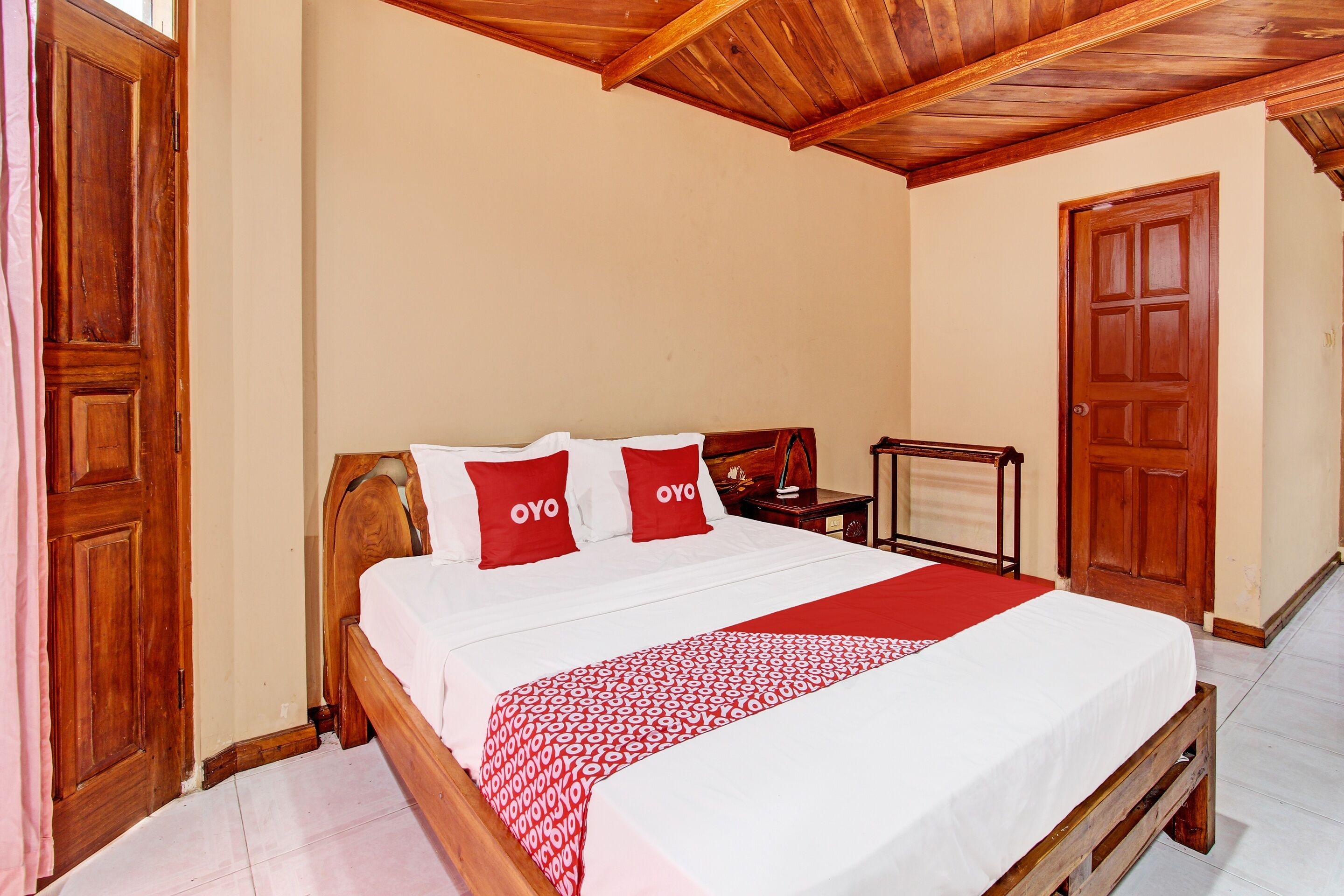 A neatly made bed with a red and white patterned blanket, two red pillows with the word "OYO" on them, wooden headboard, and a wooden nightstand with a lamp.