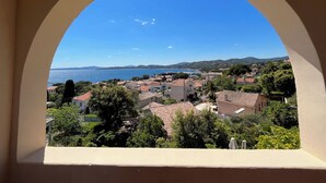 A coastal town view through an arched window, featuring houses with red-tiled roofs, greenery, and a clear blue sky.