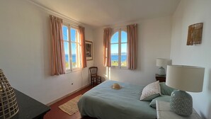 A bedroom with a bed, two windows with curtains, a chair, a lamp, and a view of the sea.