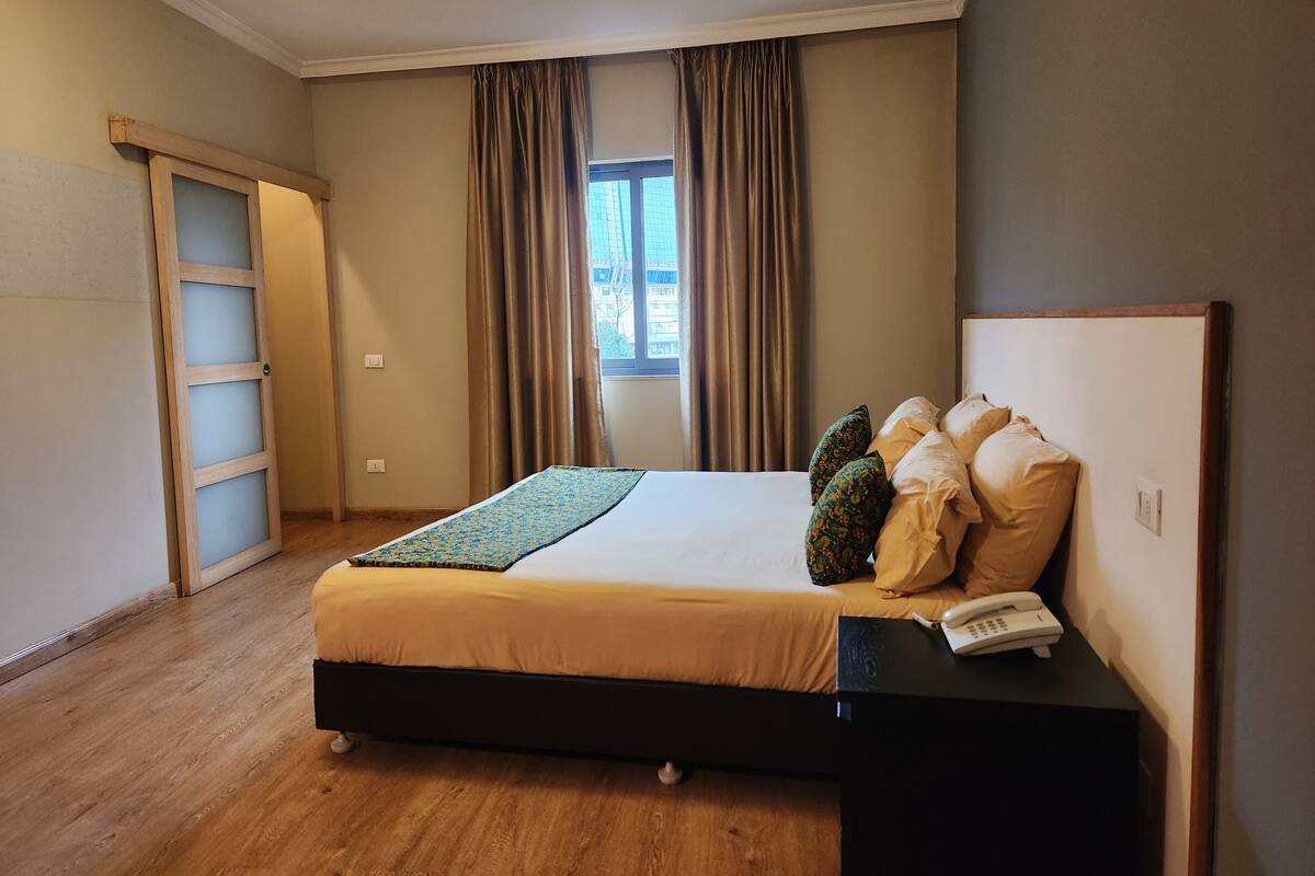 A bedroom with a bed, a bedside table with a phone, and a window with curtains.