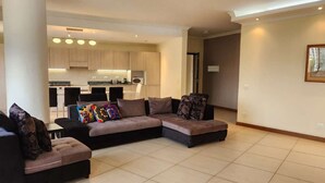 A modern living room with a sectional sofa, a small table, and a kitchen in the background. - Kora Hotel Suites (Addis Ababa)