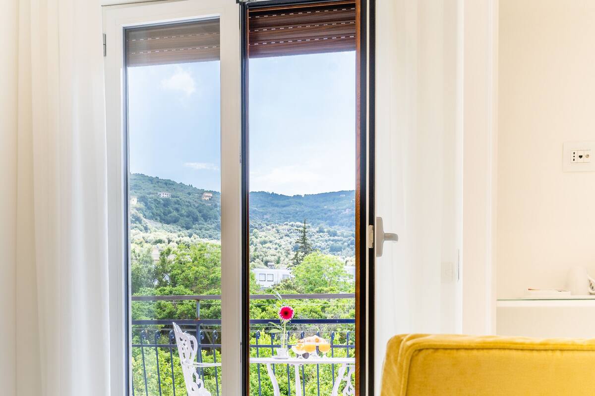 A hotel room with a bed, a yellow chair, and a balcony with a view of greenery and hills.