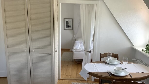 Quarto com uma cama, mesa de cabeceira com uma tigela e taça de vinho, e piso de madeira.