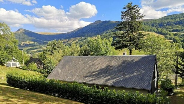 Ein Haus mit geneigtem Dach, umgeben von üppiger Vegetation und sanften Hügeln im Hintergrund.
