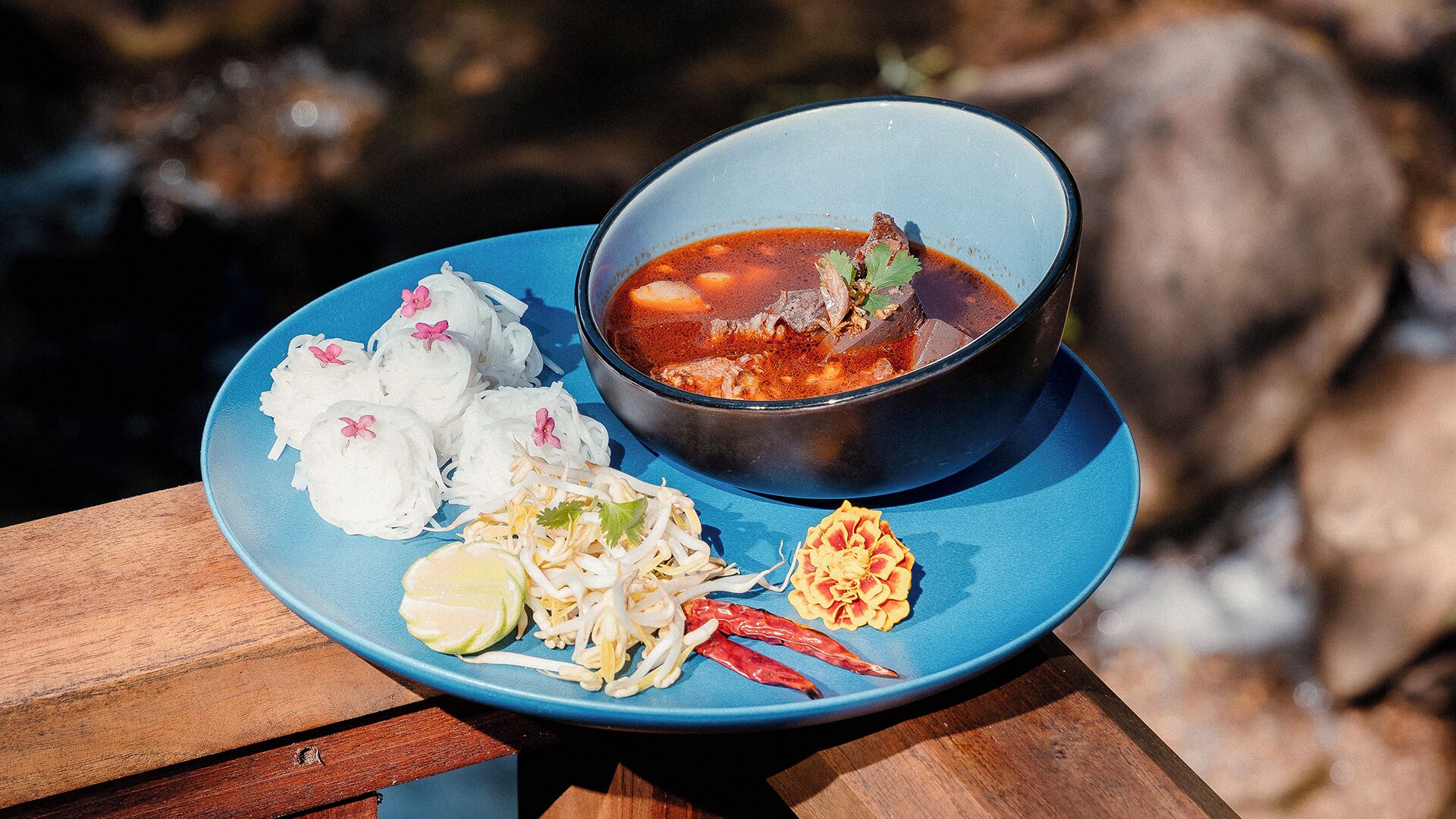 Un plato de sopa con carne y verduras, acompañado de pasteles de arroz, hierbas frescas y una guarnición de verduras encurtidas, sobre un plato azul.