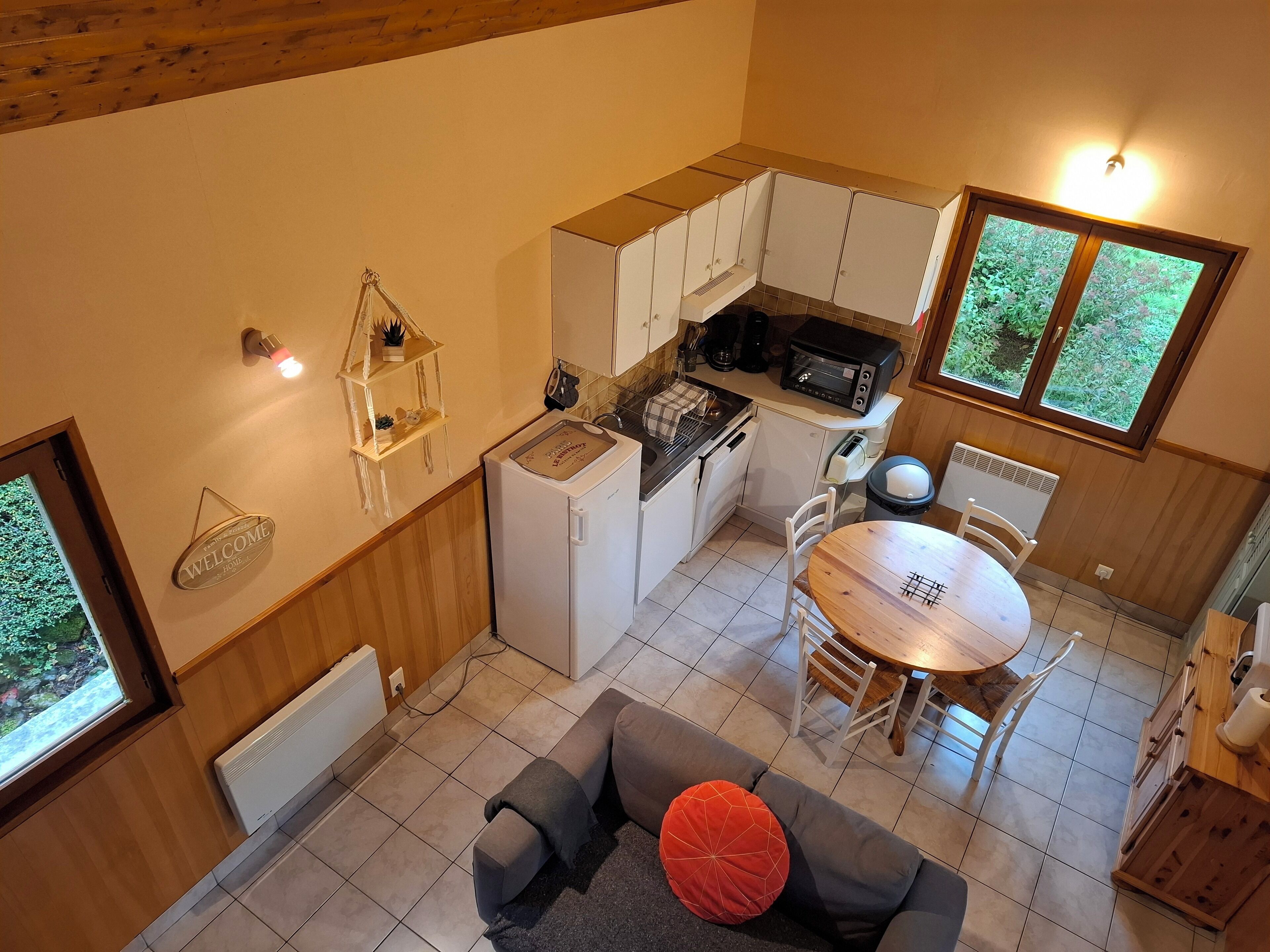 Una cocina compacta con gabinetes blancos, horno empotrado y microondas. Hay una pequeña zona de comedor con una mesa y sillas de madera. Un sofá gris con un cojín rojo está en primer plano.