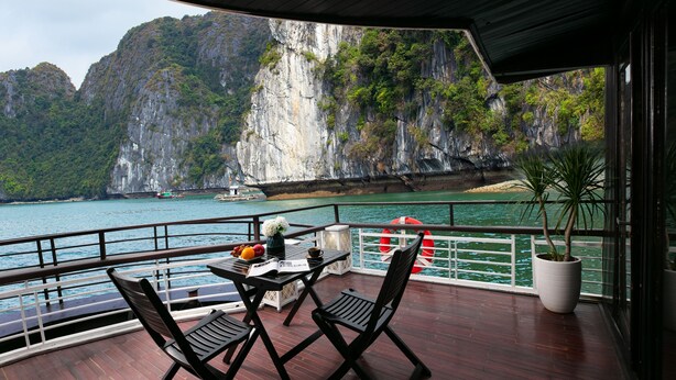 A deck with a table and chairs, overlooking a body of water with a rocky cliff in the background. - La Regina Classic Cruise (Bai Chay)