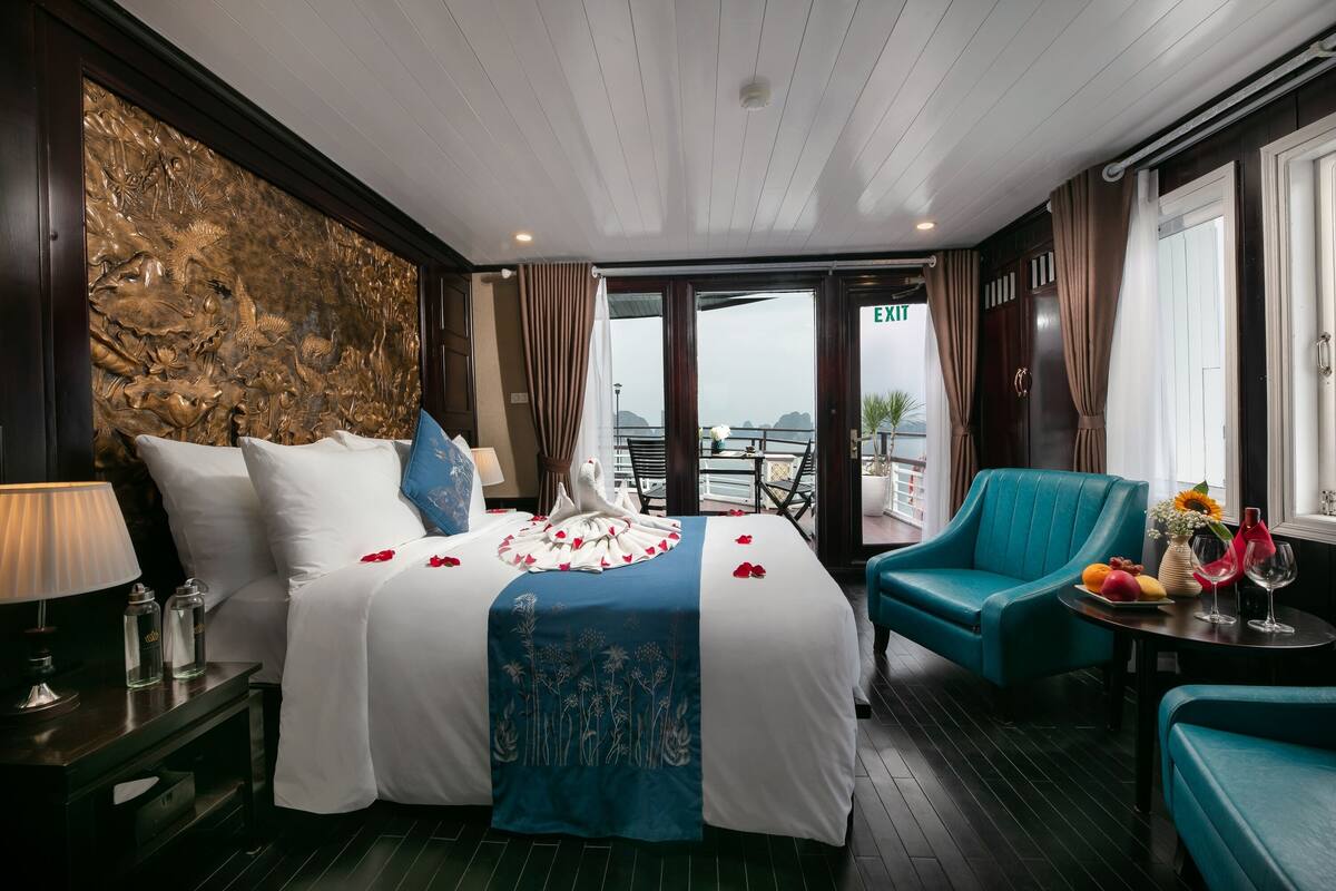 A hotel room with a bed, a blue armchair, a small table with a vase and fruit, and a view of the sea through large windows.