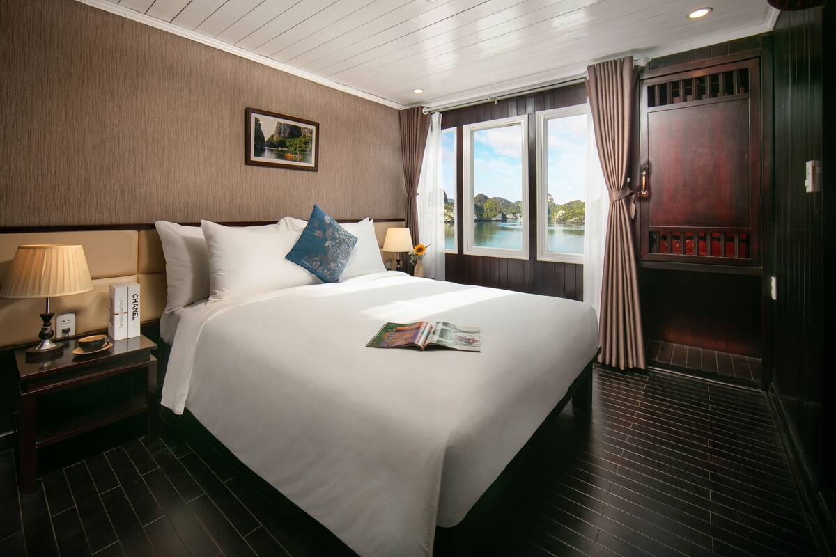 A hotel room with a large bed, a nightstand with a lamp, a window with curtains, and a view of a body of water.